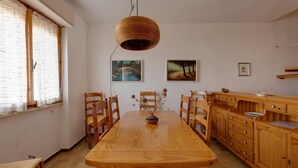 Comedor con mesa de madera, sillas y lámpara colgante. Hay dos cuadros en la pared y una ventana con cortinas.