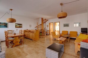 Ein großzügiger Wohnbereich mit einem Holzesstisch, Stühlen, einer Couch und einem Fernseher. Eine Treppe führt zu einer weiteren Ebene.