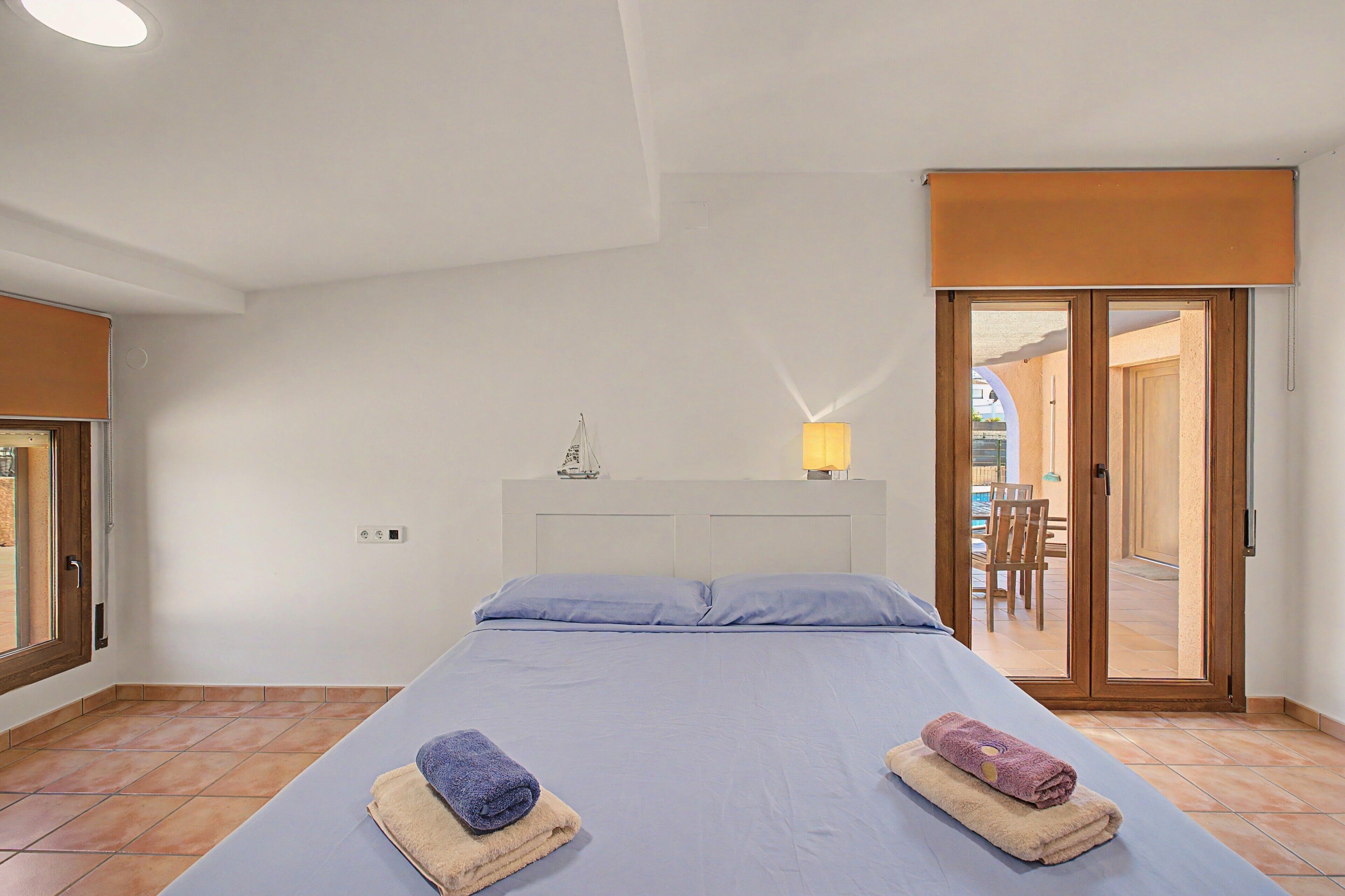 Un dormitorio con cama, dos toallas, una mesita de noche, una lámpara y vistas a un espacio exterior con mesa y sillas.