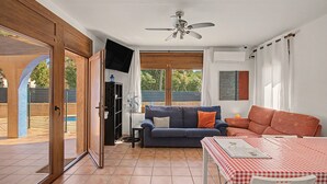 A living room with a sofa, a table with a checkered tablecloth, a ceiling fan, and a view of the outside.