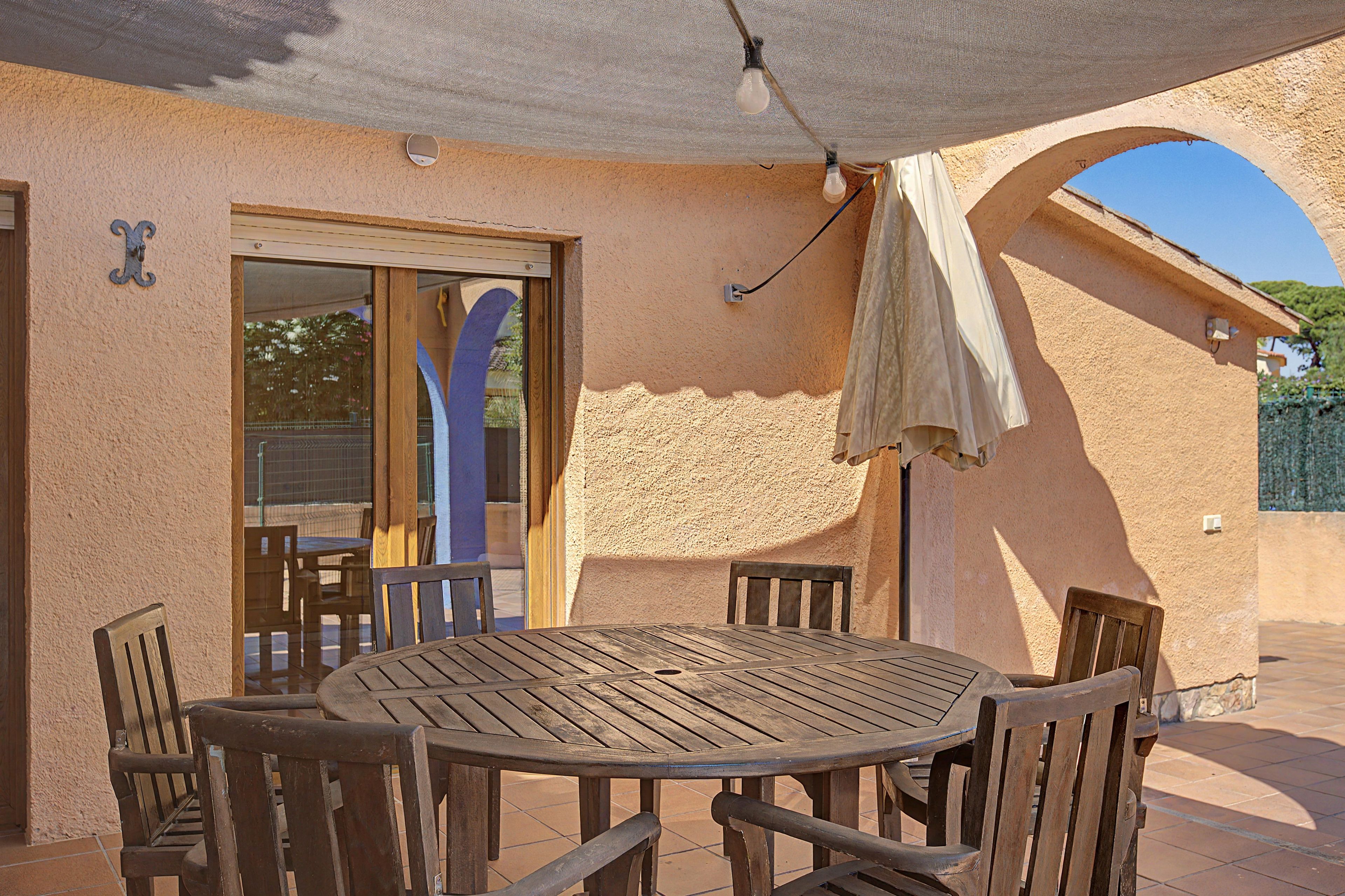 An outdoor patio with a round wooden table and chairs, a covered seating area, and a view through an archway.