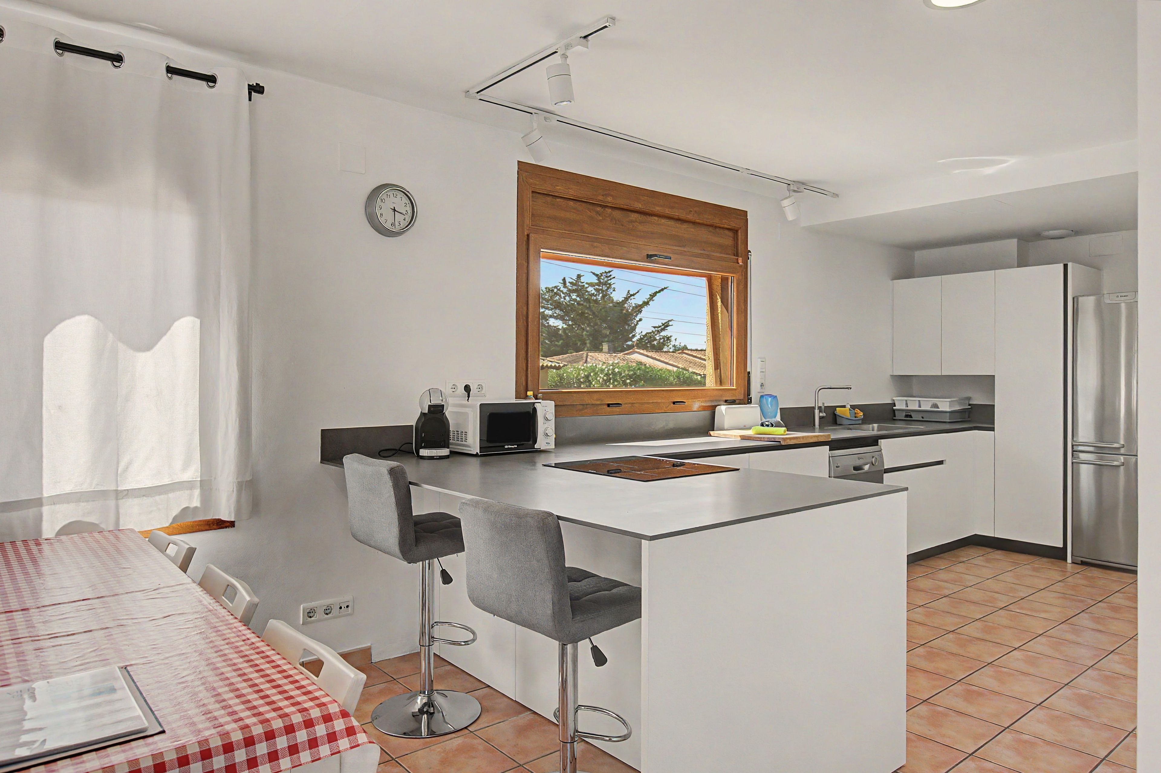 Cocina compacta con una isla integrada, taburetes de bar, una mesa de comedor con mantel a cuadros y una ventana con vistas al verdor.
