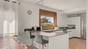 A compact kitchen with a built-in island, bar stools, a dining table with a checkered tablecloth, and a window with a view of greenery.