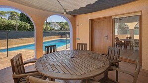 A covered outdoor seating area with a pool view, a wooden table, and chairs.