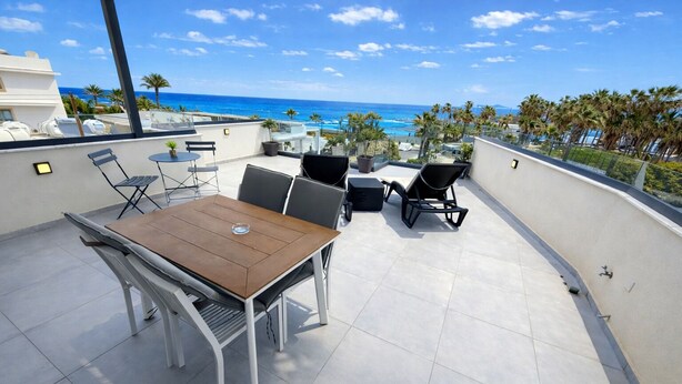 Eine Dachterrasse mit Tisch und Stühlen, mit Blick auf einen Strand und Palmen.