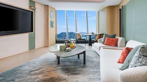 Un salón moderno con un televisor de pantalla plana grande, una mesa de centro con fruta, un sofá con cojines y vistas a montañas a través de amplios ventanales.