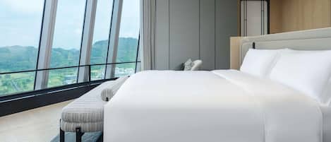 A hotel room with a large bed, a bench, and a view of a mountainous landscape through floor-to-ceiling windows.