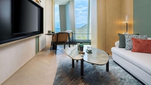 Un salón moderno con un televisor de pantalla plana, una mesa de centro redonda de vidrio, un sofá con cojines estampados y vistas al exterior a través de grandes ventanales.