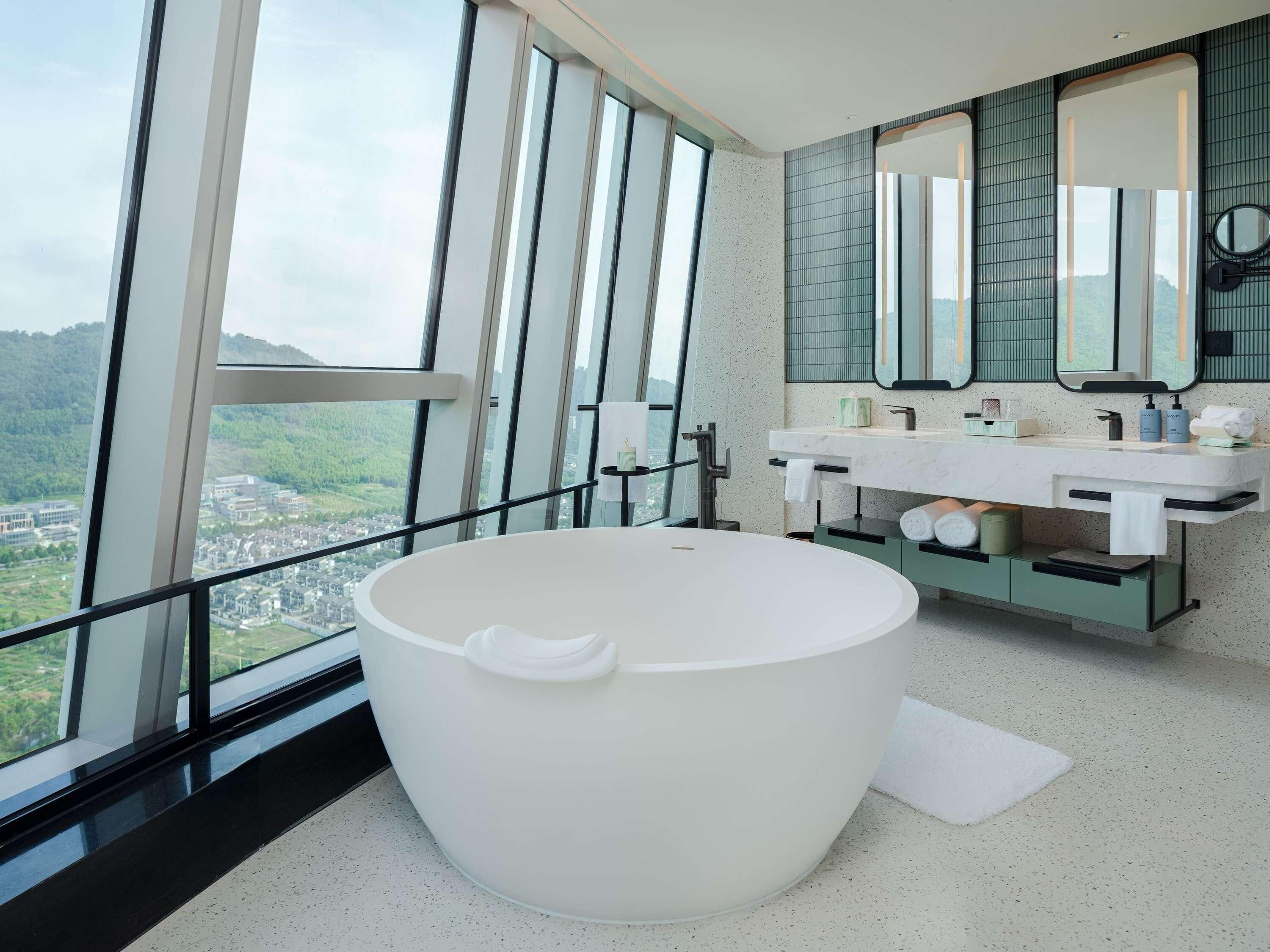 A modern bathroom with a large bathtub, a vanity with a mirror, and a view of a green landscape through large windows.