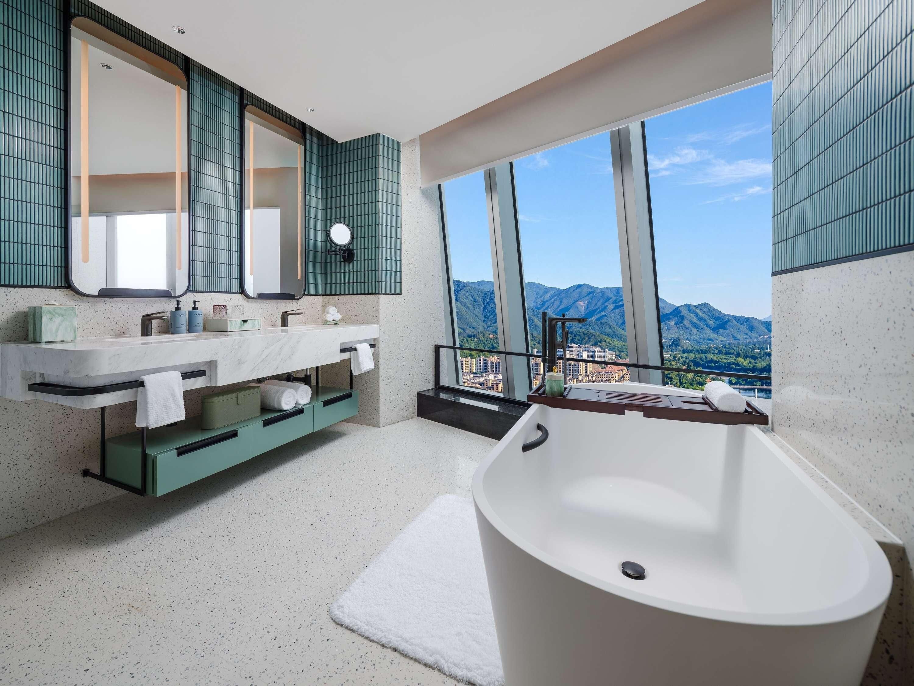 Ein modernes Badezimmer mit einer freistehenden Badewanne, einem großen Spiegel und Bergblick durch hohe Fenster.