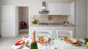 A modern kitchen with white cabinets, a stainless steel range hood, and a dining area with a table set for two. - Elegant detached house in the city center (Avola)
