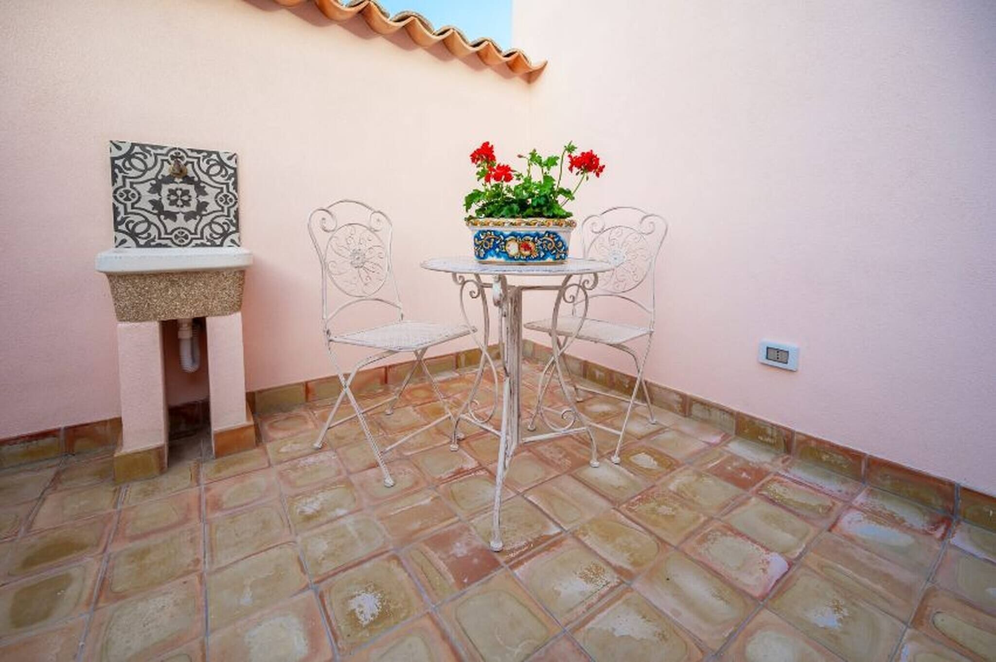 A small patio area with a tiled floor, a round table with two chairs, a decorative planter with red flowers, and a wall-mounted tile piece.