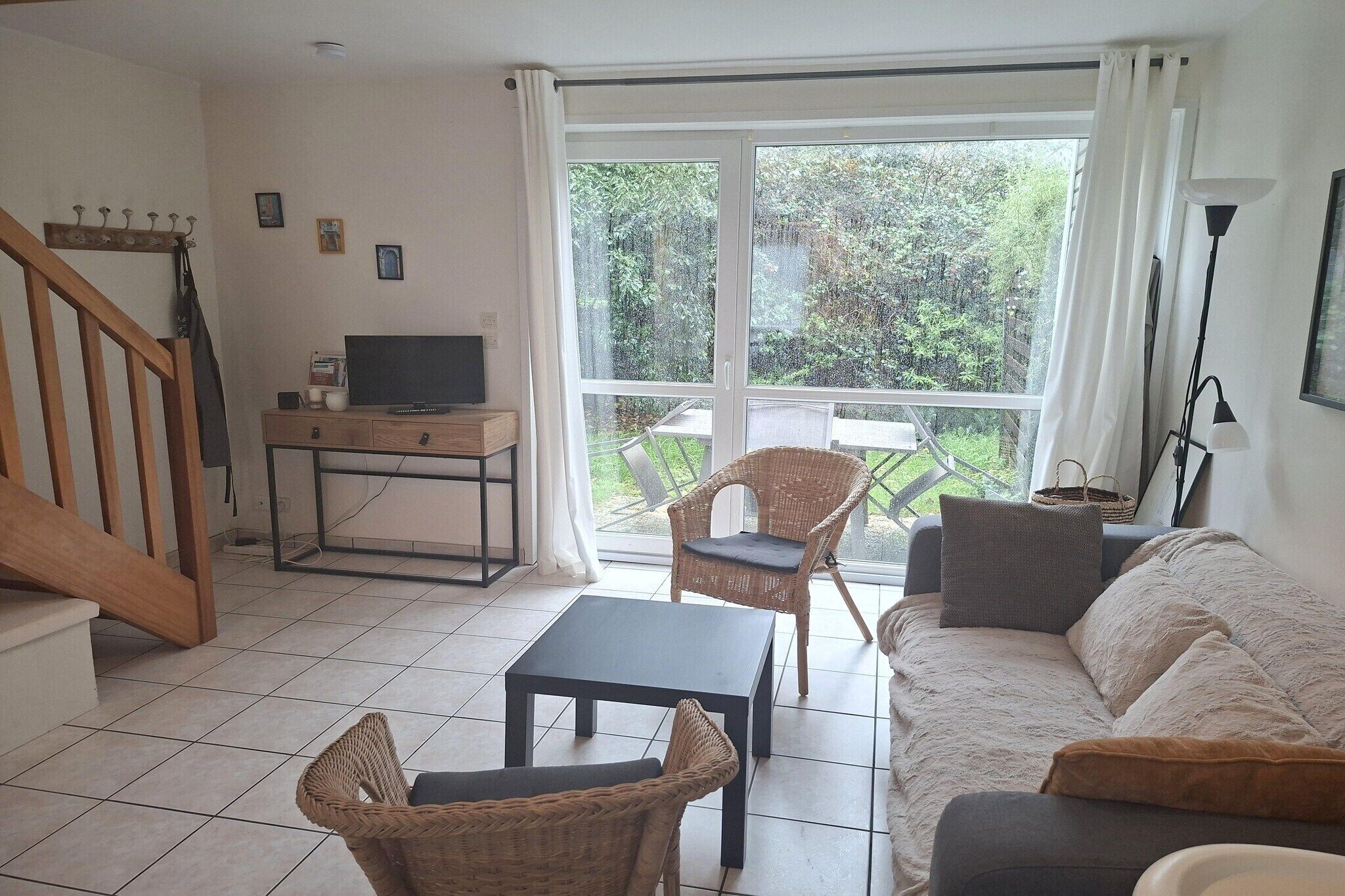A living room with a sofa, two wicker chairs, a small table, a TV on a stand, and a large window with curtains.