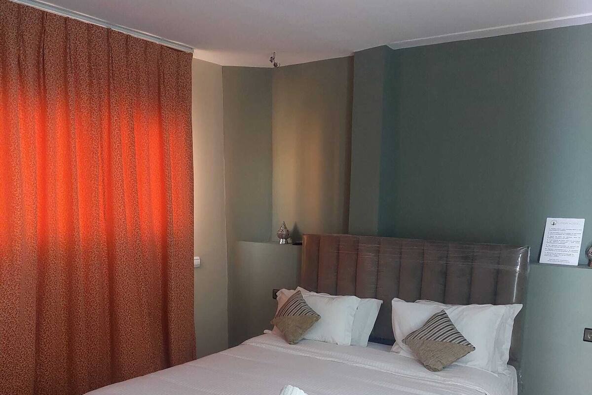 A hotel room with a large bed, a red curtain, and a headboard with multiple pillows.