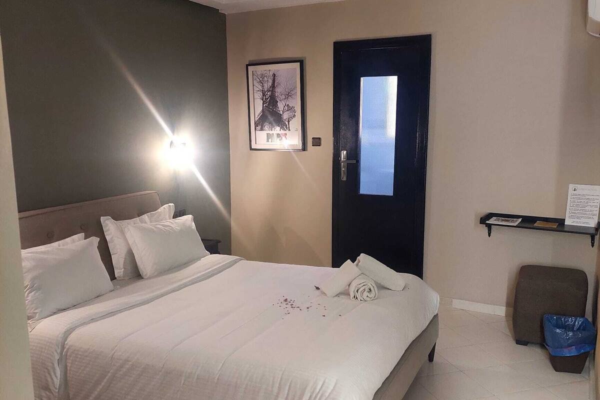 A hotel room with a bed, a door, a framed picture on the wall, and a small trash bin.