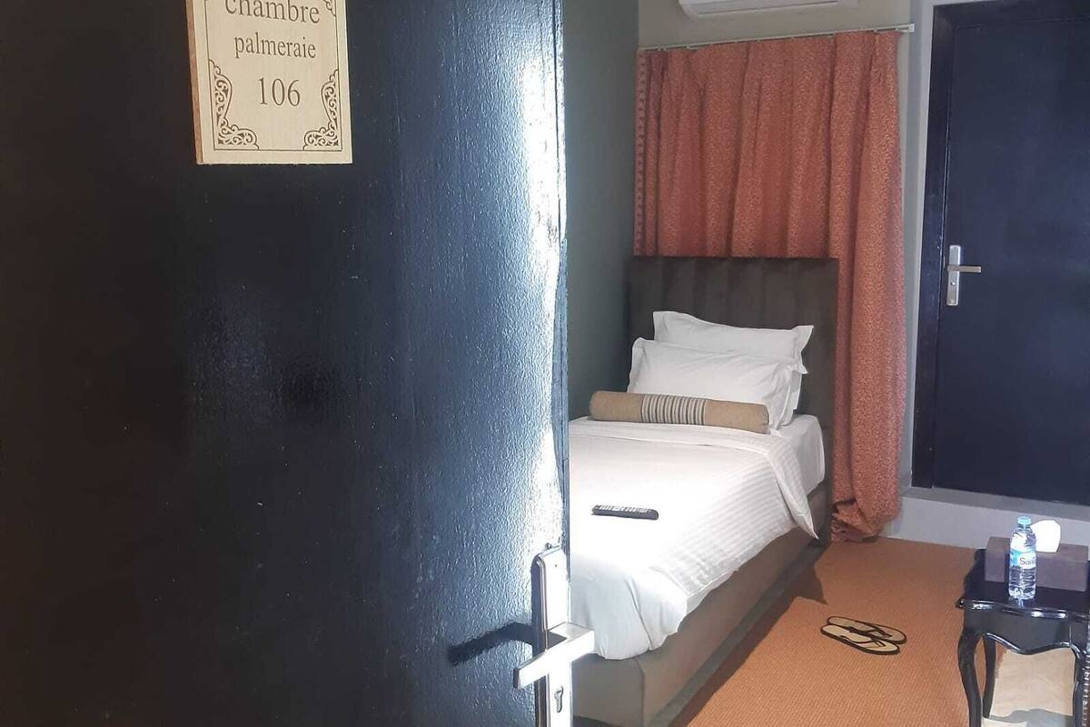 A hotel room with a bed, a bedside table with a water bottle, a door with a sign, and an air conditioning unit.