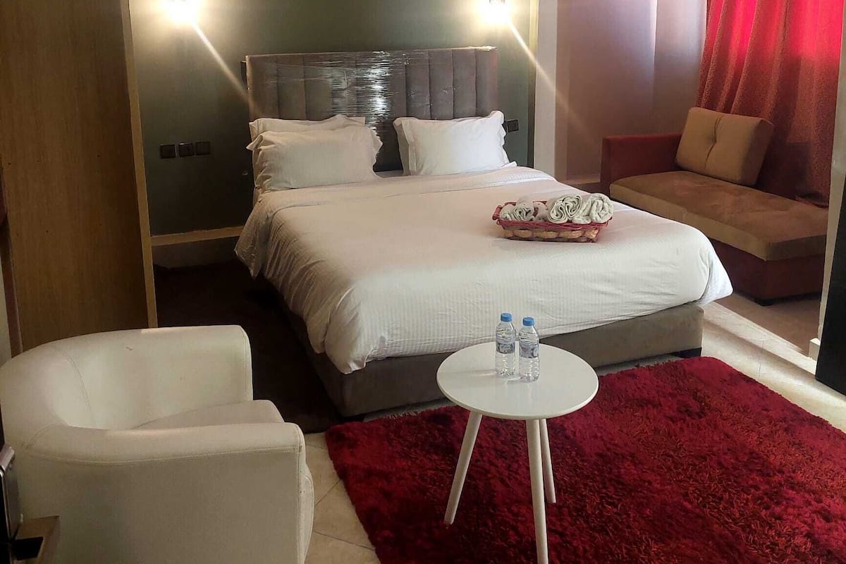A hotel room with a large bed, a small round table with two bottles, a red rug, a sofa, and a chair.