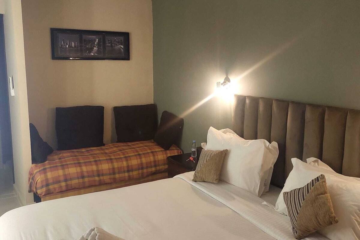 A hotel room with a large bed, a headboard with lights, a wall-mounted picture, and a nightstand with a bottle.