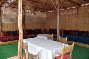 A covered outdoor seating area with woven furniture and tables, a thatched roof, and a green carpet.