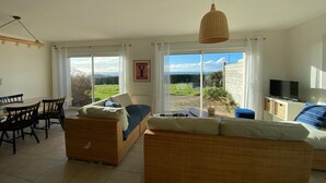 A spacious living room with a sofa, dining table, and chairs. There is a television on a stand, a wicker chair, and a large window with a view of the outdoors. - Panoramic sea view – Le Conquet (Le Conquet)