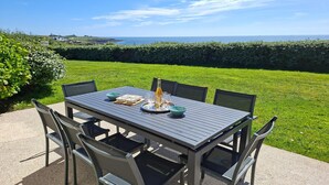 An outdoor dining table with chairs, a bottle, and glasses on a table, set against a backdrop of a grassy area and a hedge. - Panoramic sea view – Le Conquet (Le Conquet)