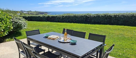 An outdoor dining table with chairs, a bottle, and glasses on a table, set against a backdrop of a grassy area and a hedge.