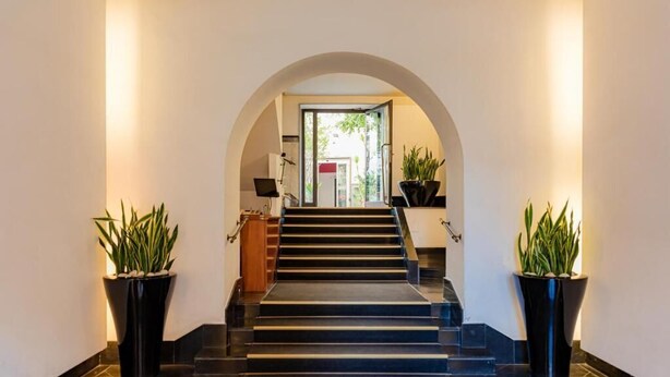 Eine moderne Hotelhalle mit einer zentralen Treppe, schwarzem Bodenbelag und an beiden Seiten Üppiges Grün in Pflanzgefäßen.