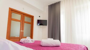 Ein Hotelzimmer mit einem Bett, einem an der Wand montierten Fernseher, einem Holzschrank mit Milchglas-Türen und einem Fenster mit grauen Vorhängen.