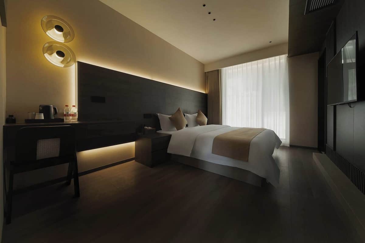 Een moderne hotelkamer met een groot bed, nachtkastjes, een bureau met stoel, een flatscreen televisie en een groot raam met gordijnen.