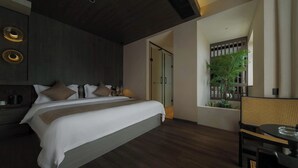 Une chambre d’hôtel moderne avec un grand lit, une table de chevet avec une lampe, une petite table avec un verre, et une vue sur un balcon agrémenté de plantes.