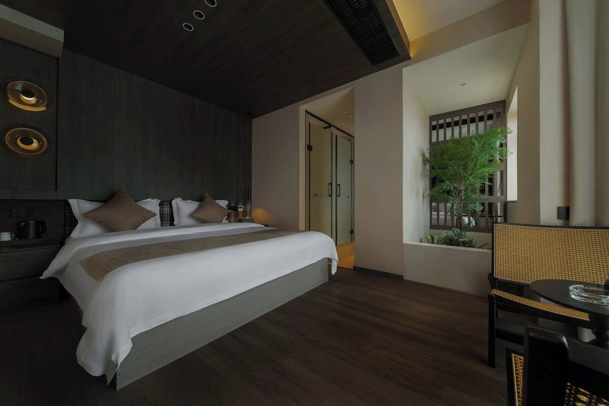 Een moderne hotelkamer met een groot bed, een nachtkastje met een lamp, een kleine tafel met een glas, en uitzicht op een balkon met planten.