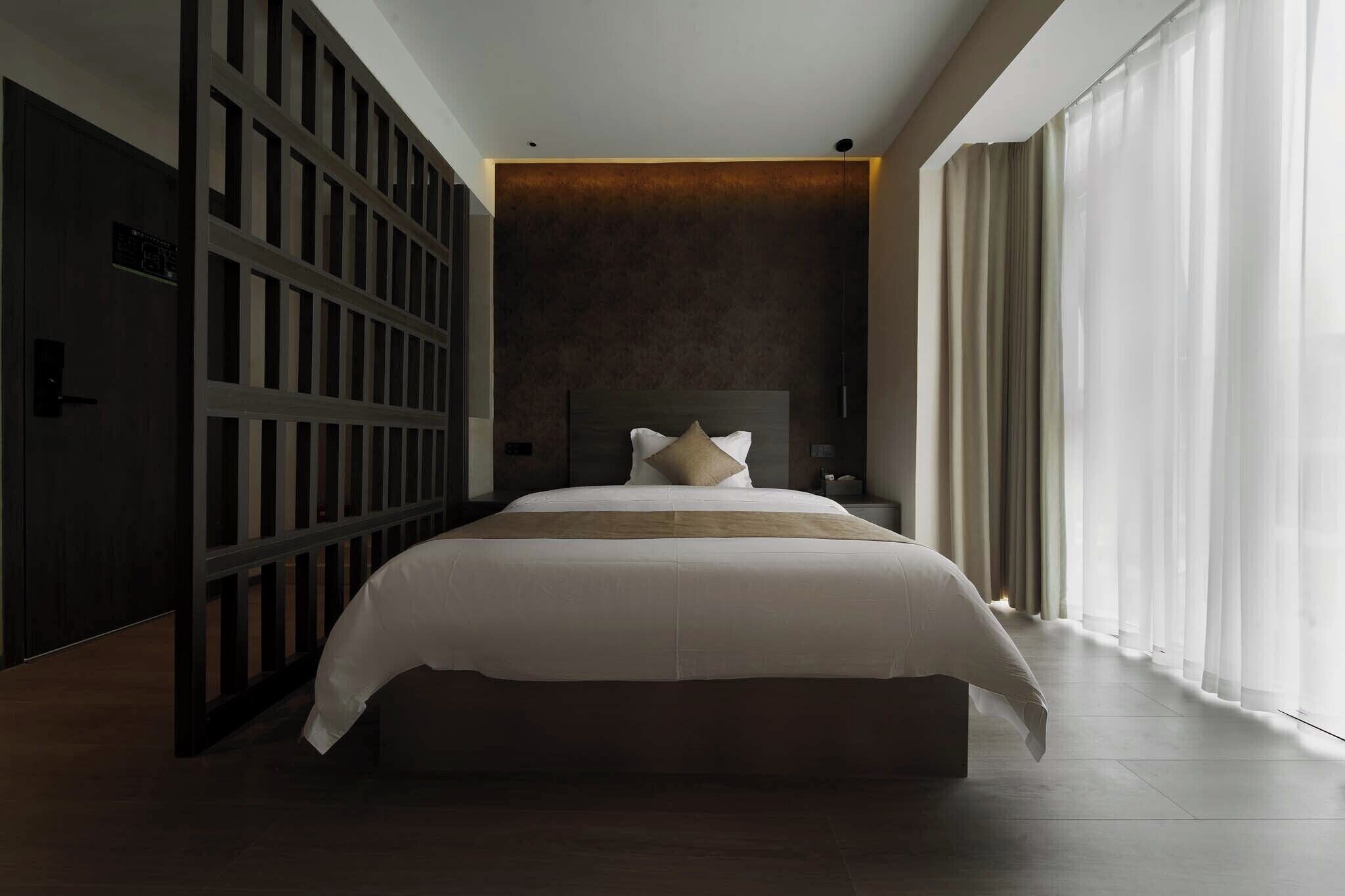 Een moderne hotelkamer met een groot bed, een donkere scheidingswand en een groot raam met dunne gordijnen.
