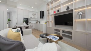 Ein modernes Wohnzimmer mit Sofa, Couchtisch und Fernseher. Im Hintergrund befindet sich eine Küche mit Schränken und einem Essbereich.