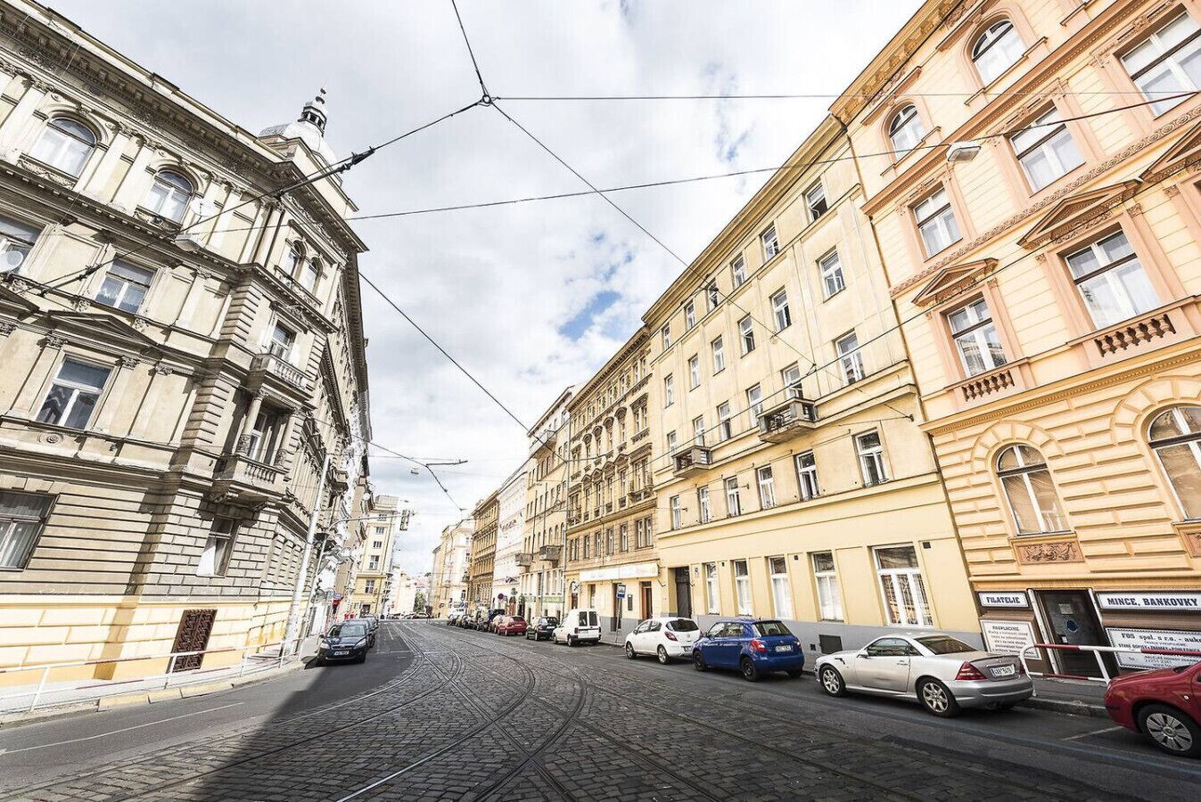 A cobblestone street lined with historic buildings, a tram track, and parked cars.