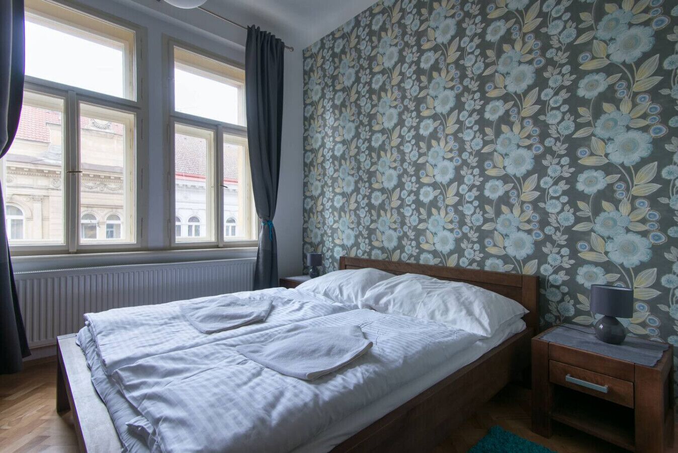 Une chambre à coucher avec un lit, une table de chevet, une fenêtre avec des rideaux et un papier peint à motifs floraux.