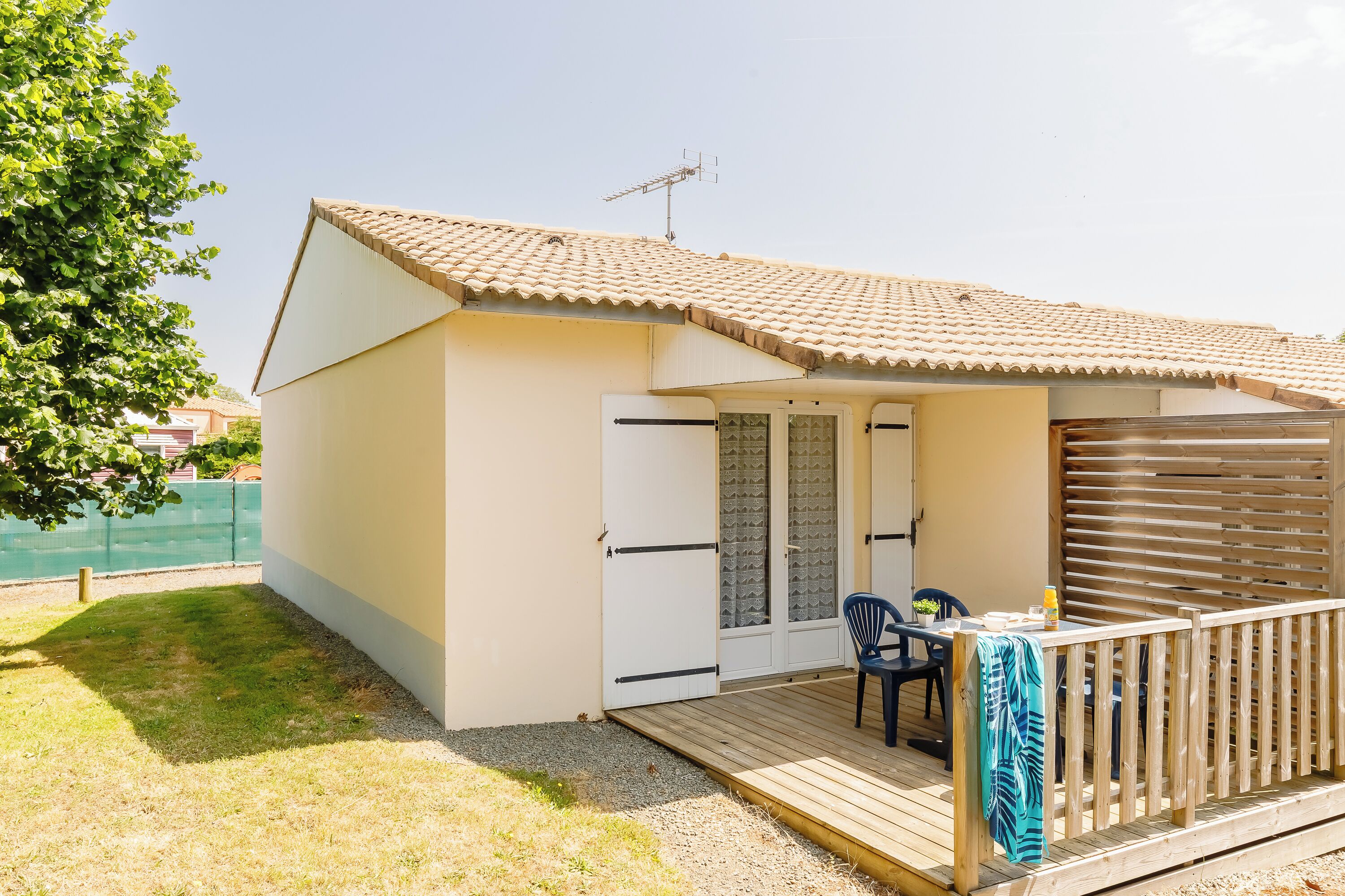 Un petit bâtiment d’un seul étage, doté d’un toit en tuiles, d’une terrasse en bois et d’un espace extérieur abrité avec une table et des chaises.