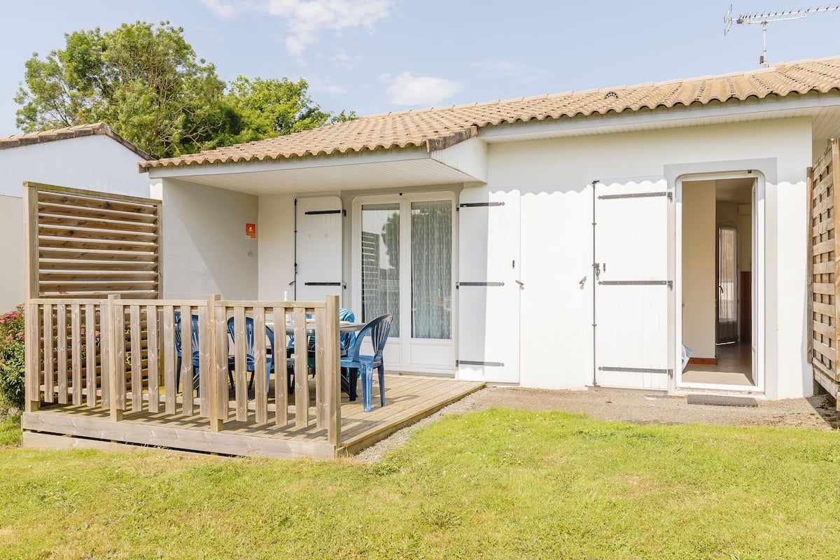 A white building with a tiled roof, a wooden deck with chairs, and a sliding glass door.