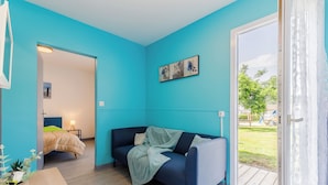 A modern room with a teal wall, a dark blue sofa, a wooden table, and a view of a playground through a glass door. - Village Vacances Gîtes Les Rivières (Maché)