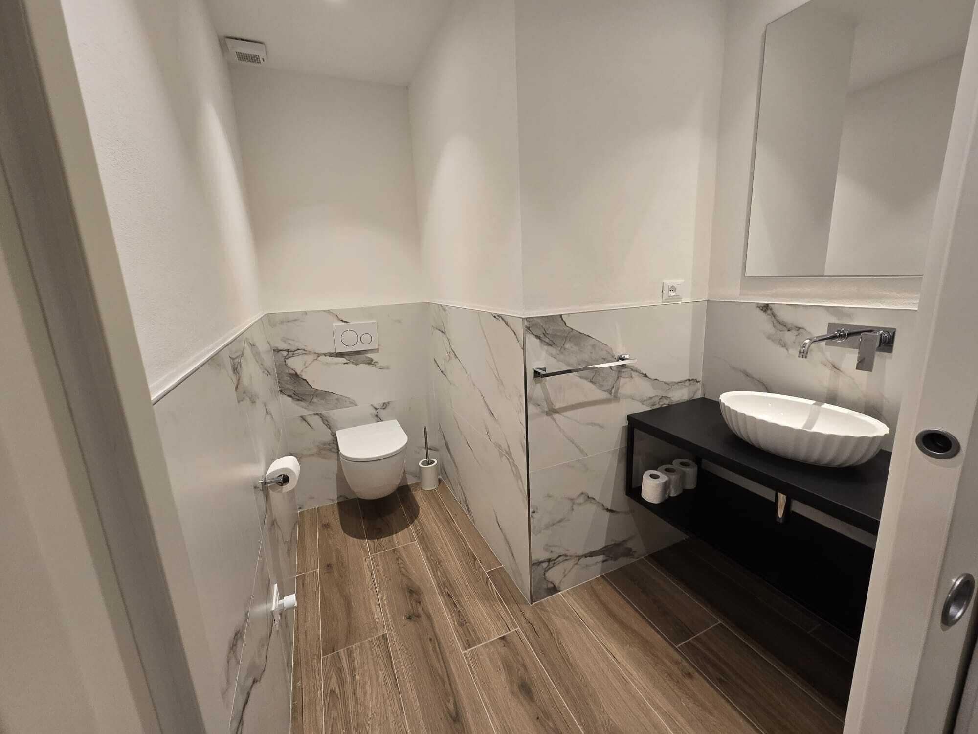 Ein modernes Badezimmer mit Marmorwand, Holzboden, weißer Toilette und einem schwarzen Waschtisch mit weißem Becken.