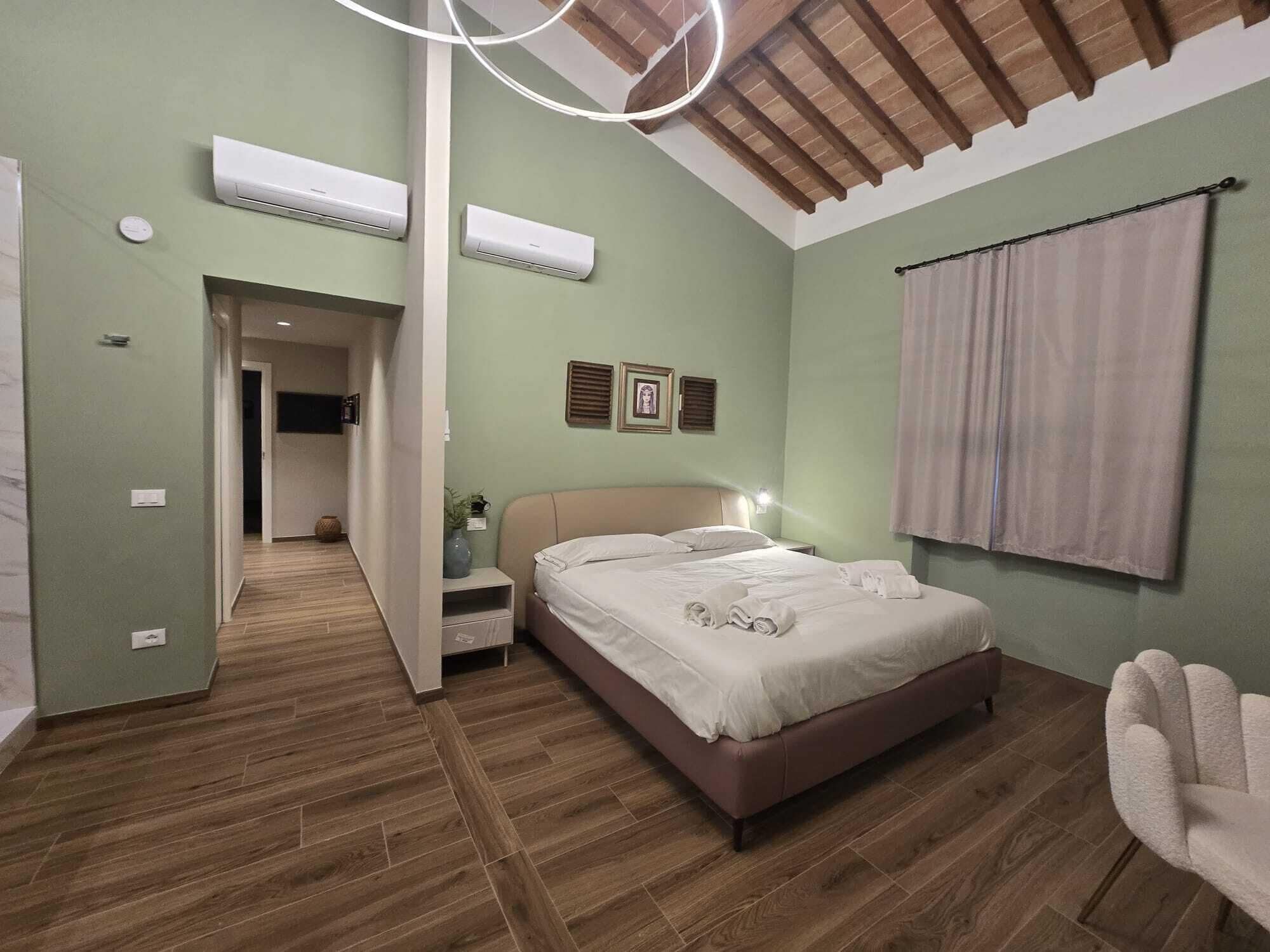 A bedroom with a bed, bedside tables, a nightstand, a wall-mounted air conditioner, a ceiling fan, and a window with curtains.