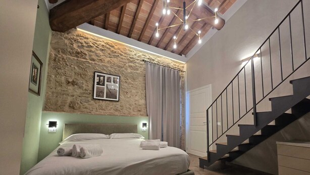Une chambre avec un mur en pierre, un lit recouvert de draps blancs, un escalier et un luminaire au plafond.