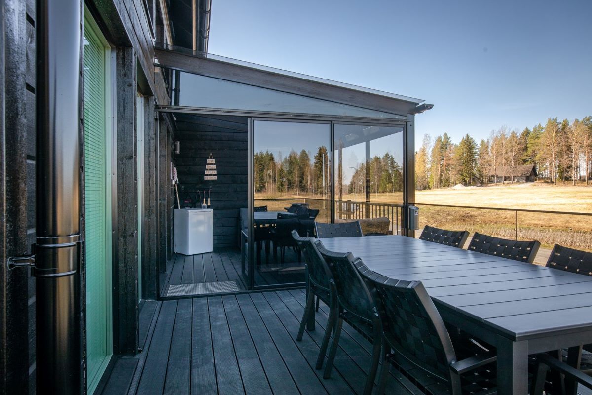 A wooden deck with a table and chairs, a glass door leading to an outdoor area, and a view of trees and a field.