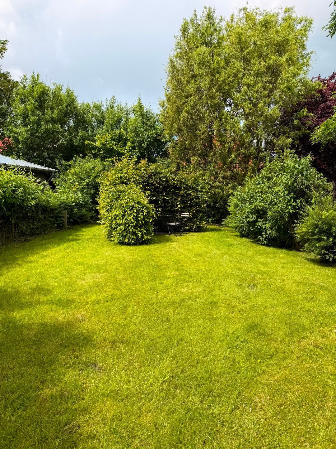 A well-maintained grassy lawn with various shrubs and trees, a wooden fence, and a metal roof structure.