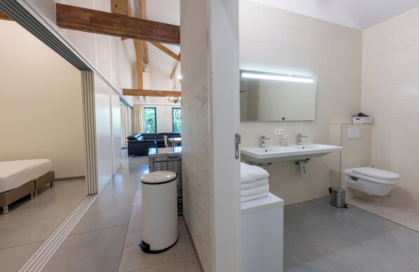 A modern bathroom with a large mirror, a sink, and a toilet. There is a bench in the adjacent room.