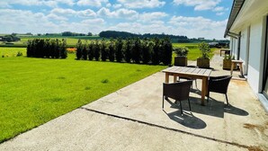 Un patio con tavolo e sedie, pavimento in cemento e vista su un campo verde e alberi.