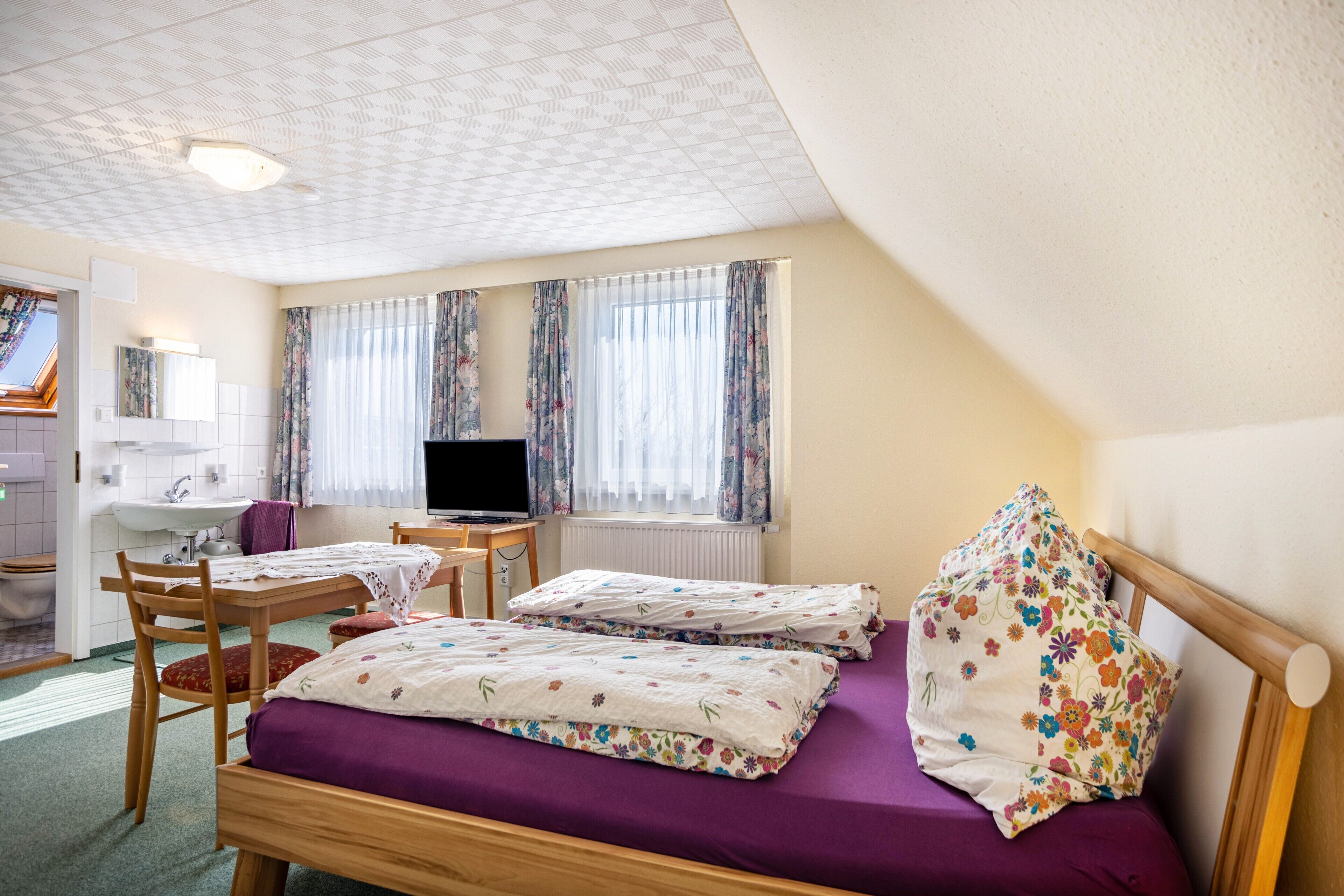 A bedroom with two beds, a desk, a chair, a television, and a window with curtains.