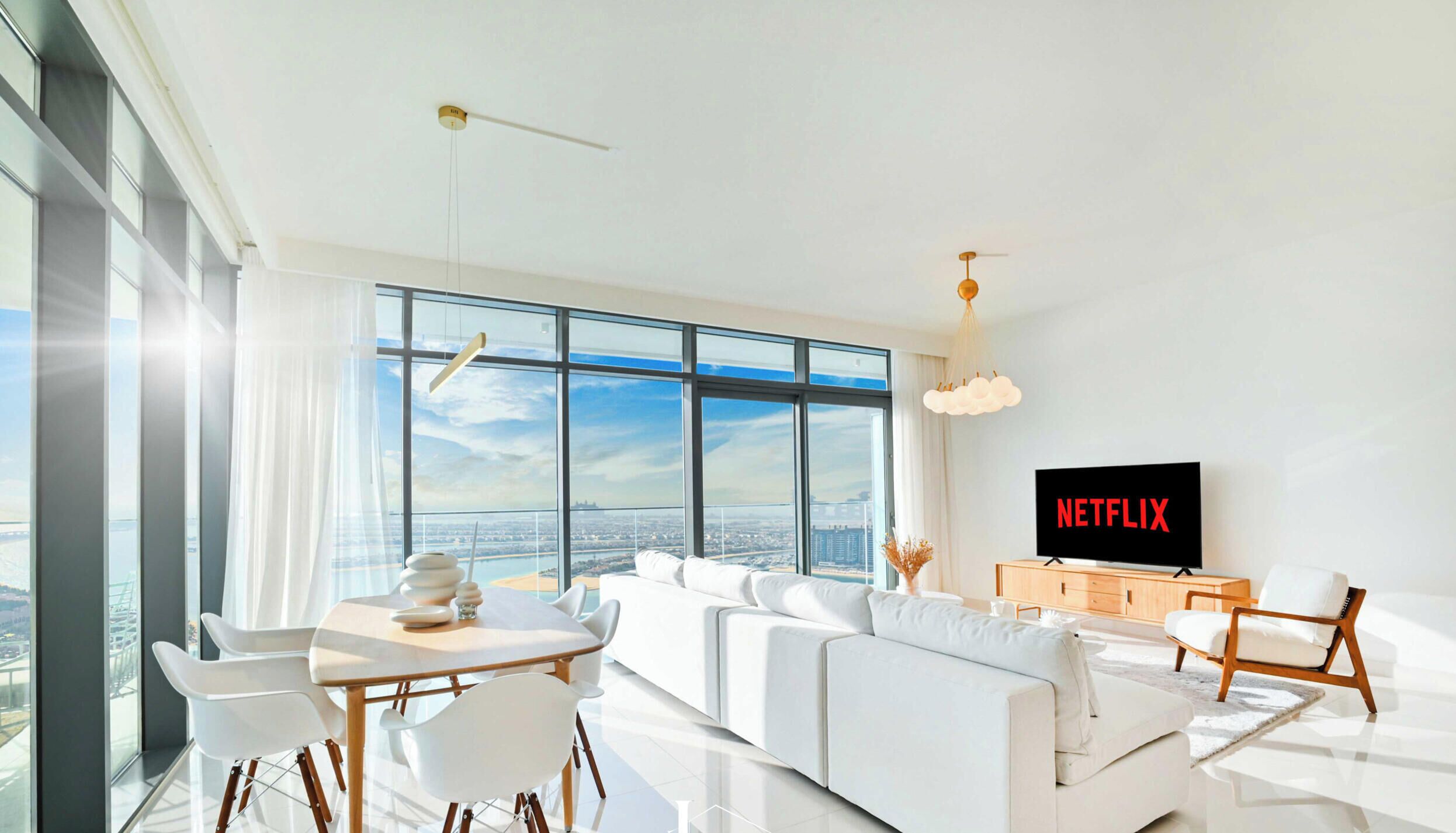 Un soggiorno moderno con una grande finestra, arredi bianchi, una TV con Netflix e vista sulla città.