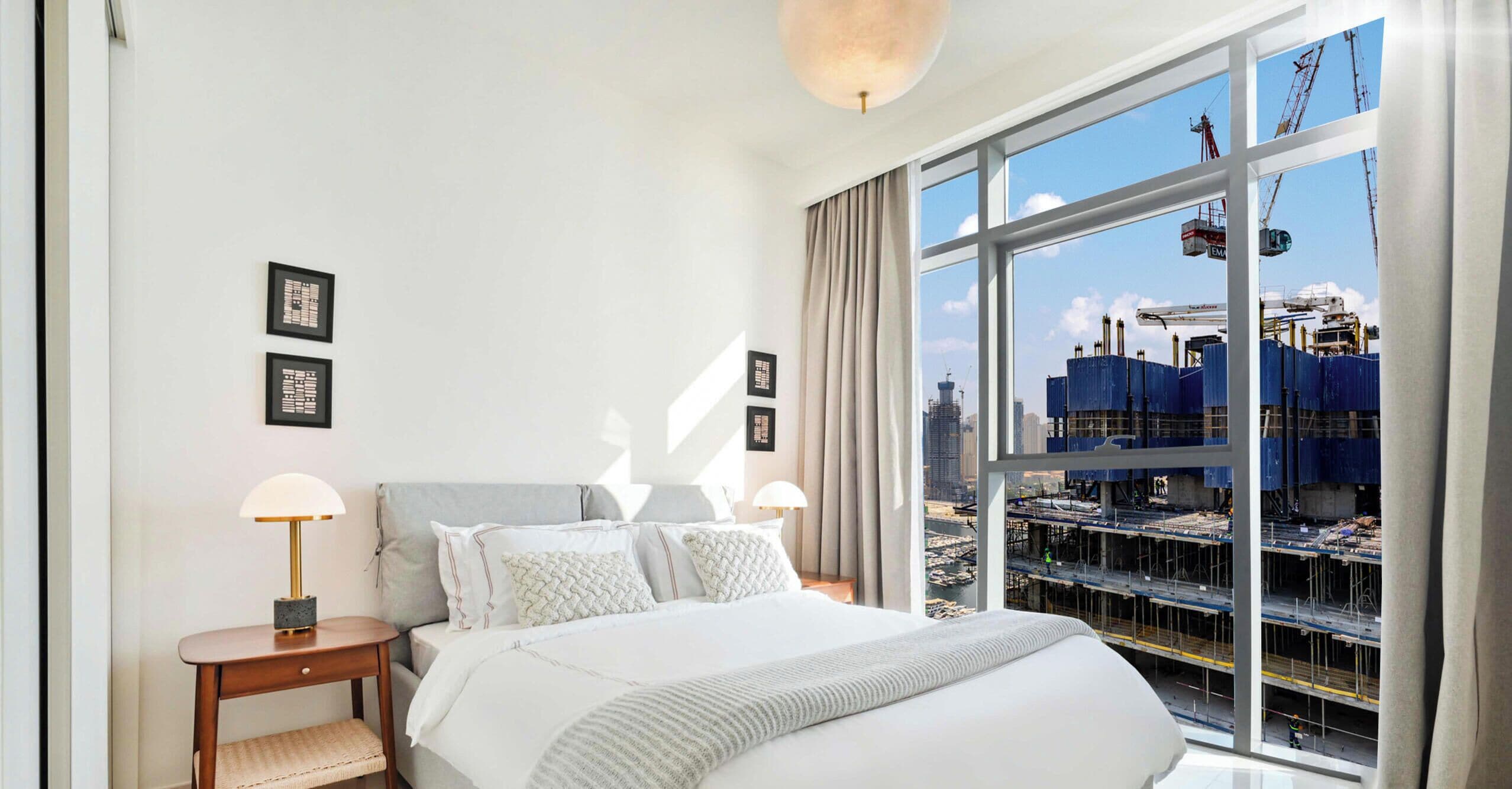 Een hotelkamer met een groot raam, een bed, een nachtkastje en kunstwerken in lijsten aan de muur.
