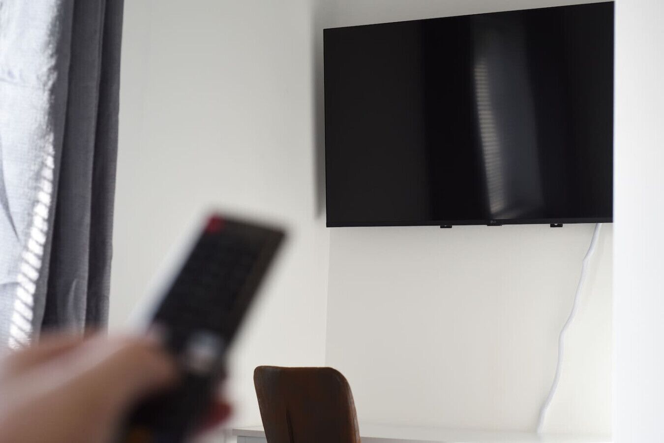 A hand holding a remote control in front of a television mounted on a wall.