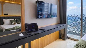 Moderni hotellihuone, jossa on suuri televisio, työpöytä kannettavalla tietokoneella, sänky ja parveke, jolta on näkymä.